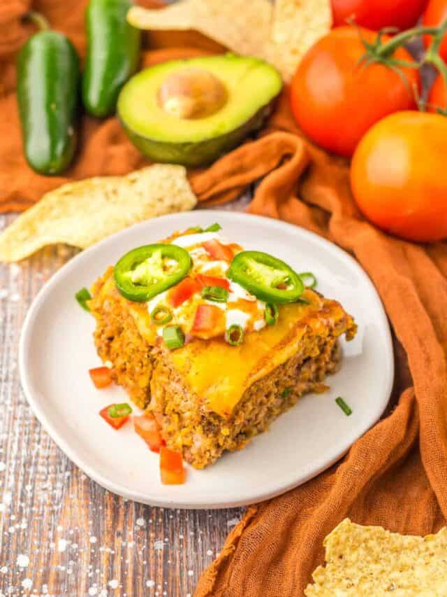 A slice of casserole topped with cheese, sour cream, diced tomatoes, and jalapeños on a white plate. Surrounding the plate are jalapeños, an avocado, tomatoes, and tortilla chips.