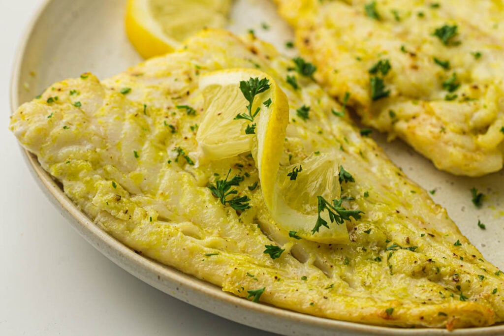 Air fryer cod fillet cooked to perfection, garnished with fresh lemon slices and chopped parsley on a beige plate.