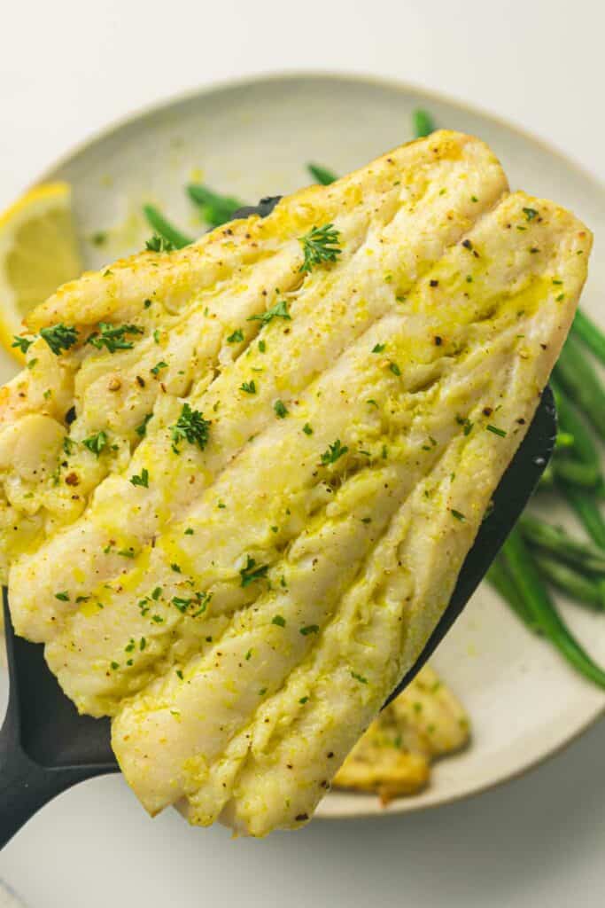 A close-up of a piece of air fryer cod garnished with herbs, held by a spatula. In the background, a plate with green beans and a lemon wedge is partially visible.