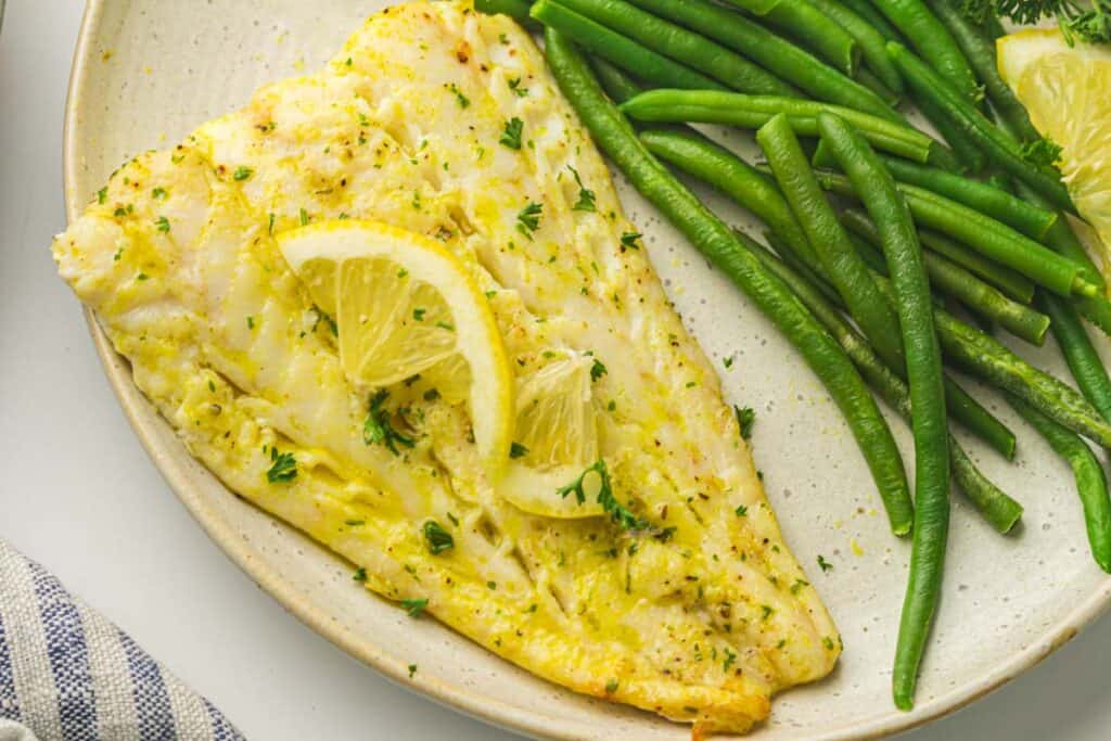 A plate with air fryer cod fillet topped with lemon slices and garnished with herbs, served alongside steamed green beans.