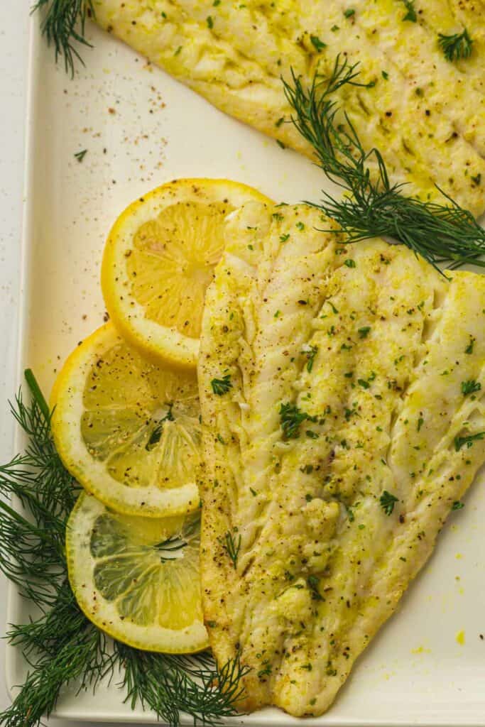 Grilled cod fillets seasoned with herbs, garnished with lemon slices and fresh dill on a white plate. Enjoy the same delicious flavor by preparing these fillets in an air fryer for a healthier twist.