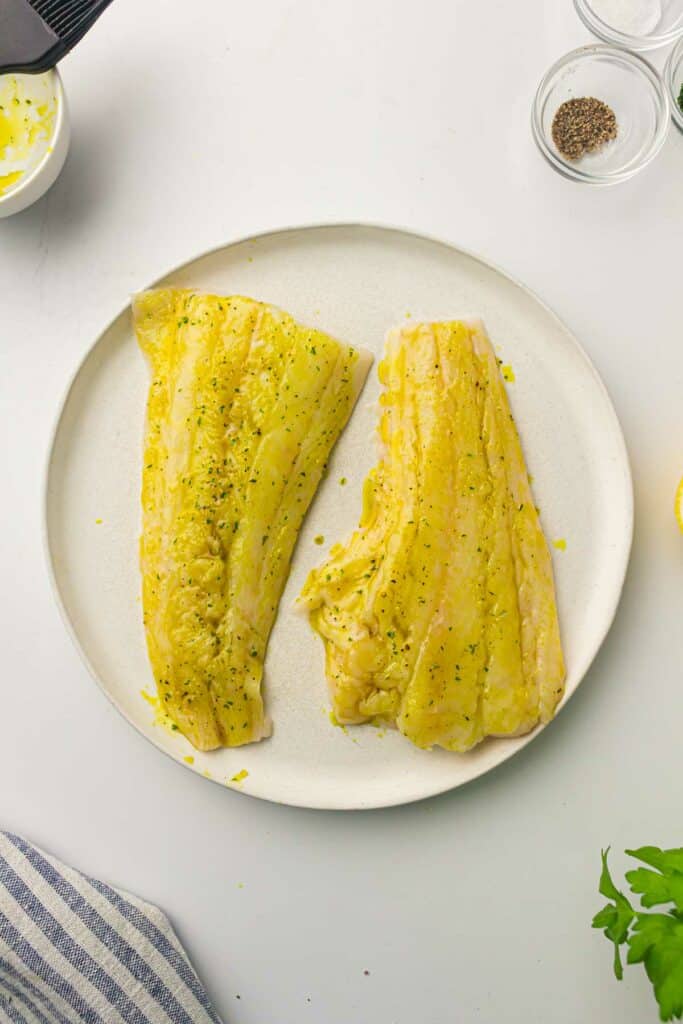 Two uncooked fish fillets on a white plate, marinated with yellow seasoning and sprinkled with herbs, sit ready for the air fryer. Beside them is a small bowl of pepper and other ingredients.