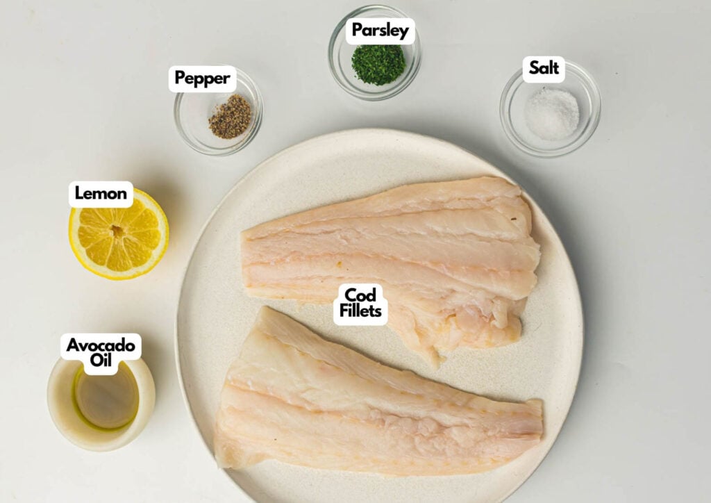 Two raw cod fillets on a plate, surrounded by small bowls of avocado oil, black pepper, parsley, salt, and a halved lemon—perfect for preparing air fryer cod. Each ingredient is clearly labeled for easy cooking.