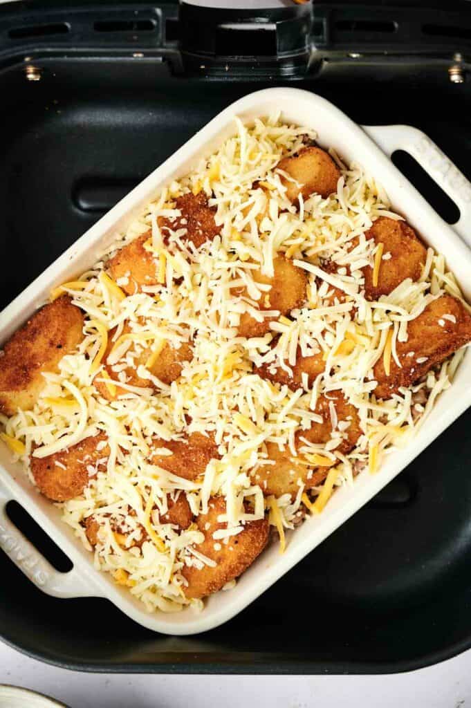 A white baking dish filled with breaded and fried rolls, sprinkled with shredded cheese, sits inside a black air fryer like a delicious twist on an air fryer tater tot casserole.