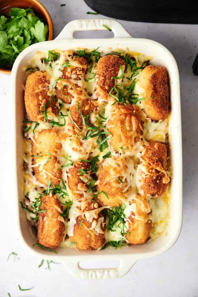 A rectangular baking dish filled with a golden, cheesy tater tot casserole topped with crispy breaded rolls and garnished with chopped herbs.