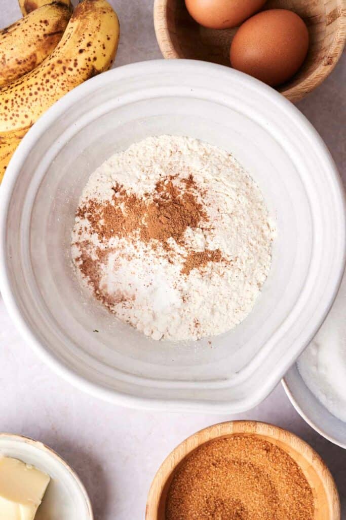 Top view of a white bowl containing flour and cinnamon, surrounded by ingredients including bananas, eggs, butter, and brown sugar, hinting at delicious banana bread preparations.