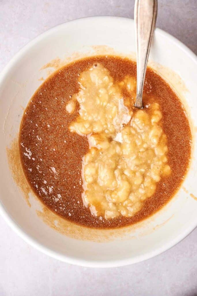 A bowl containing a mix of brown liquid and mashed bananas, reminiscent of the early stages of banana bread batter, with a spoon immersed in the mixture.