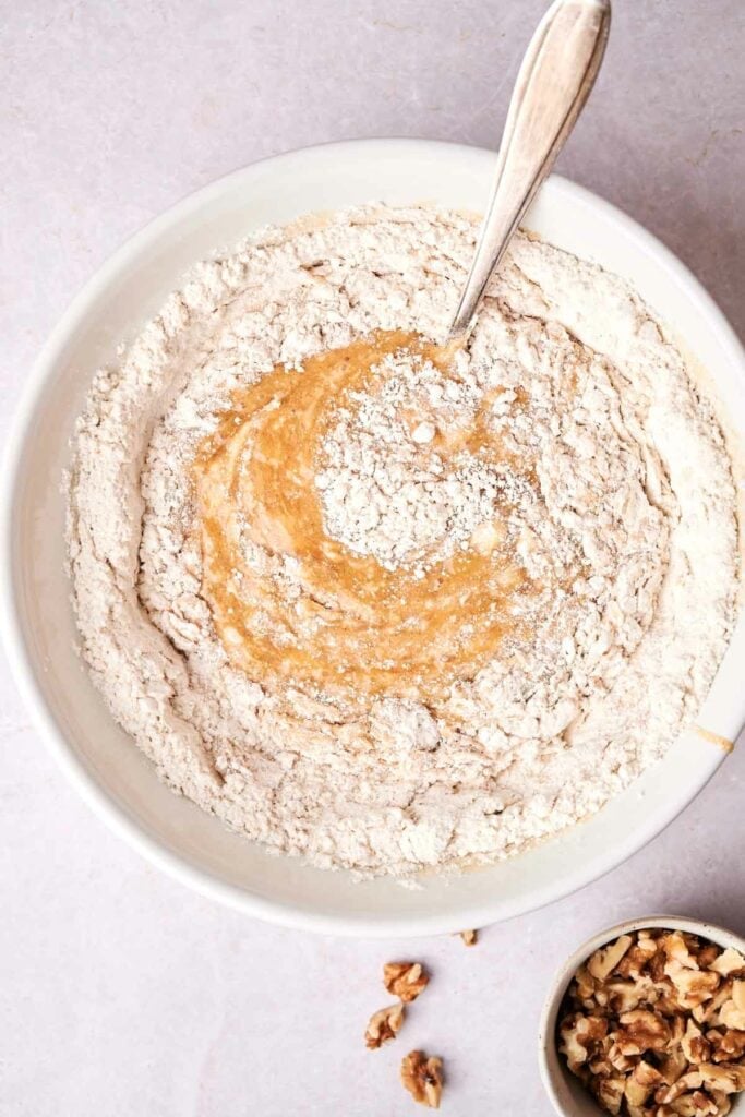 A bowl with a flour mixture and a spoon stirring in light brown banana bread batter. A small container of chopped walnuts is partially visible at the bottom.