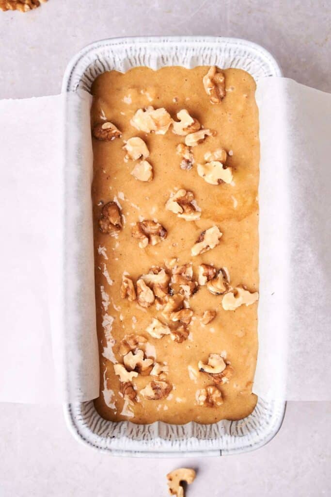 A rectangular cake pan lined with parchment paper is filled with unbaked banana bread batter topped with walnut pieces.