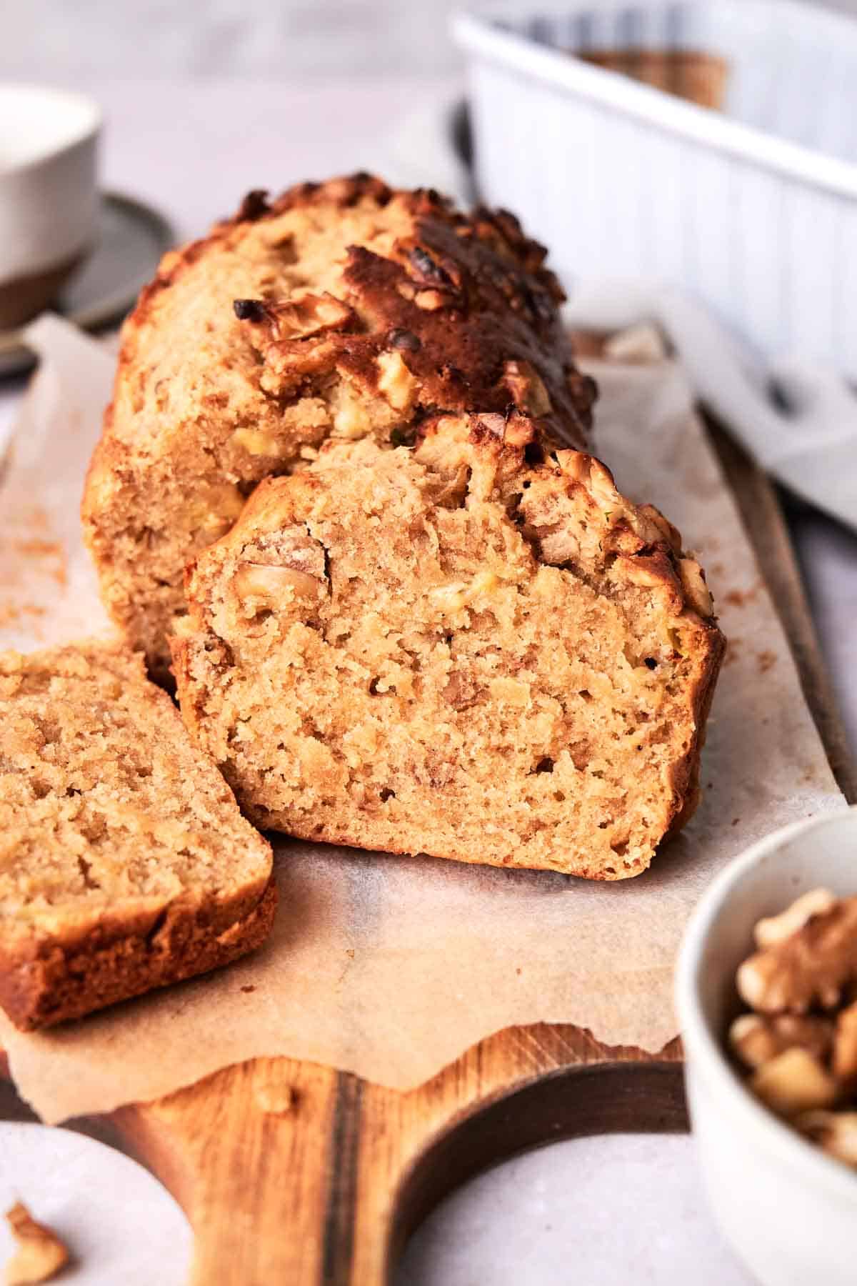 Sliced loaf of banana bread with nuts on a wooden cutting board, with a bowl of nuts and a white baking dish in the background.