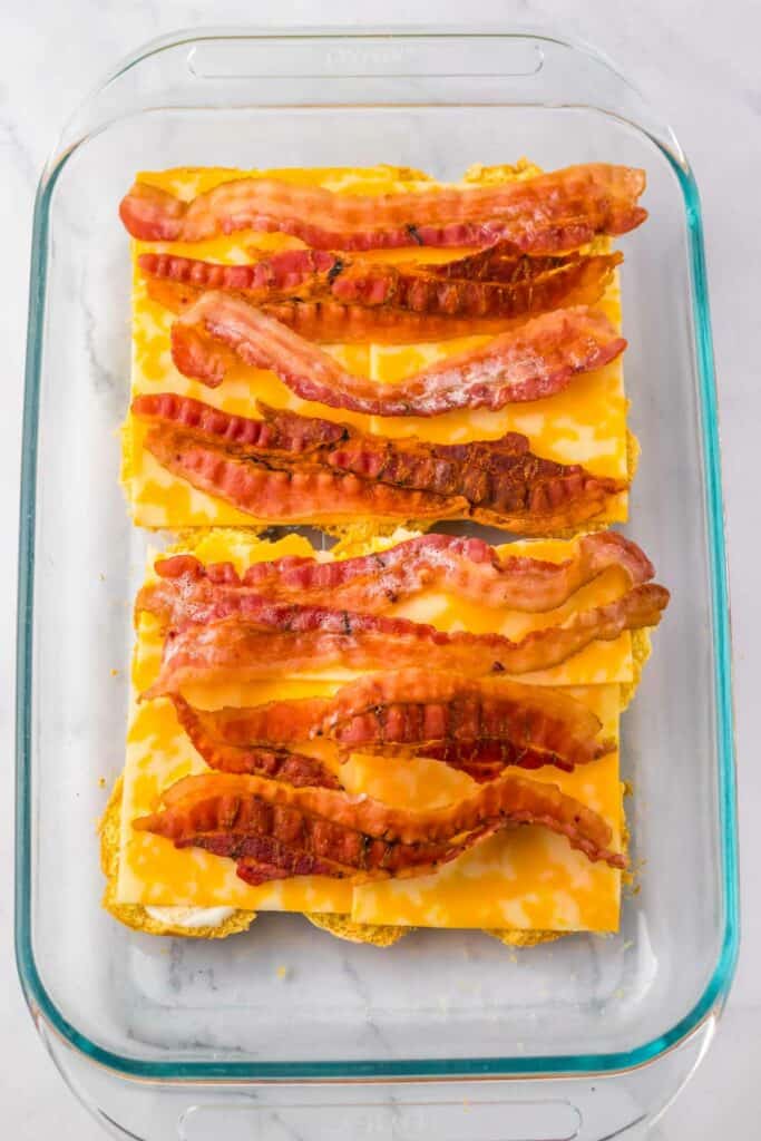 A glass dish with layers of cheese slices, cooked bacon strips, and tender chunks of chicken, perfect for making chicken bacon ranch sliders.