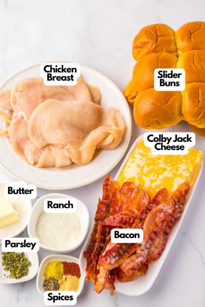 Ingredients for chicken bacon ranch sliders displayed on a table include raw chicken breast, slider buns, Colby Jack cheese, cooked bacon, butter, ranch dressing, parsley, and spices.