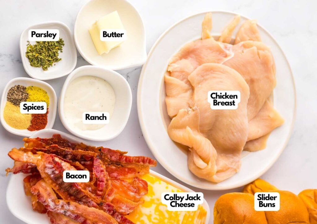 Top-down view of labeled ingredients for chicken bacon ranch sliders, featuring chicken breast, slider buns, Colby Jack cheese, crispy bacon, ranch dressing, butter, parsley, and assorted spices.