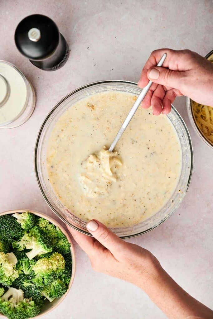 Two hands hold and stir a bowl filled with a creamy mixture, with broccoli florets and cooked chicken in a separate bowl at the bottom left, hinting at a delicious chicken and broccoli casserole. In the top left background, there's a pepper grinder and cream container.