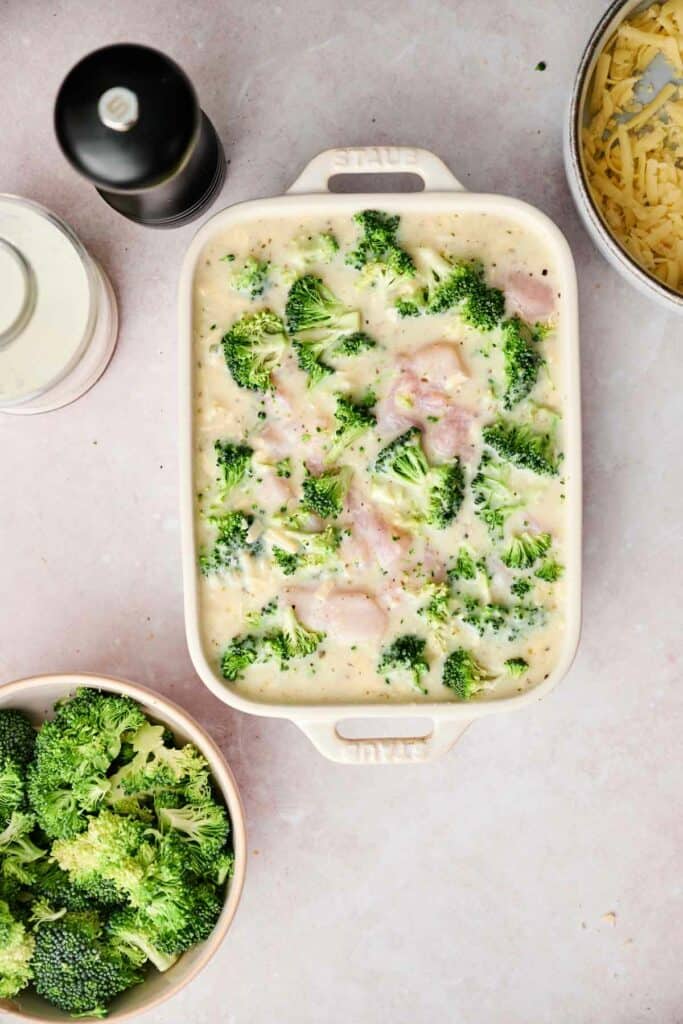 A chicken and broccoli casserole dish filled with a creamy mixture of chopped chicken, broccoli, and cheese sits on the countertop, alongside a bowl of raw broccoli florets, a bowl of shredded cheese, and a pepper grinder.
