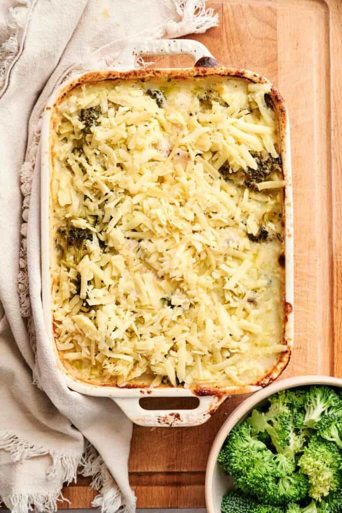 A casserole dish filled with a baked chicken and broccoli casserole, featuring a golden, melted cheese topping. A bowl of fresh broccoli florets is placed beside the dish on a wooden surface.