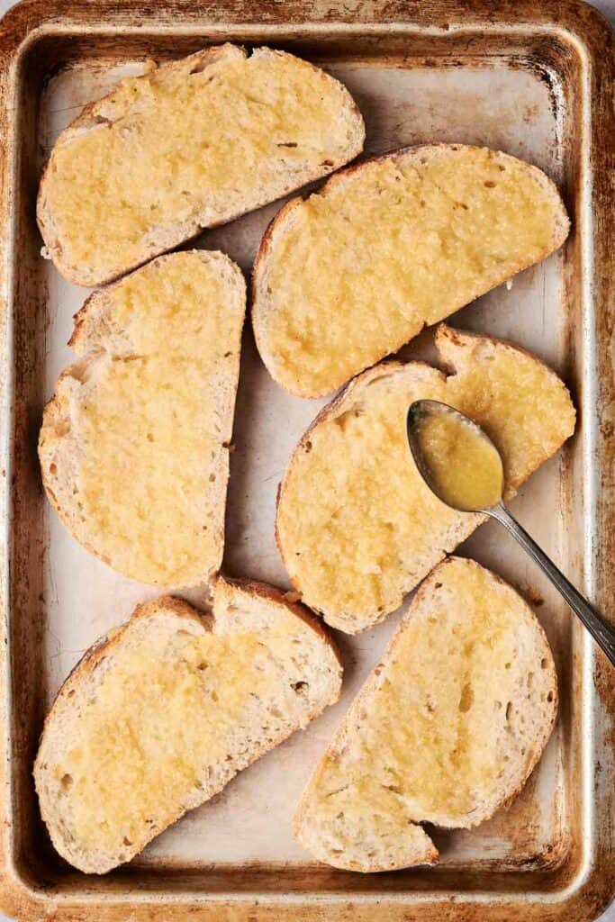 Slices of bread spread with butter on a baking sheet, with a spoon placed on one slice, transform into garlic bread by adding a sprinkle of minced garlic.