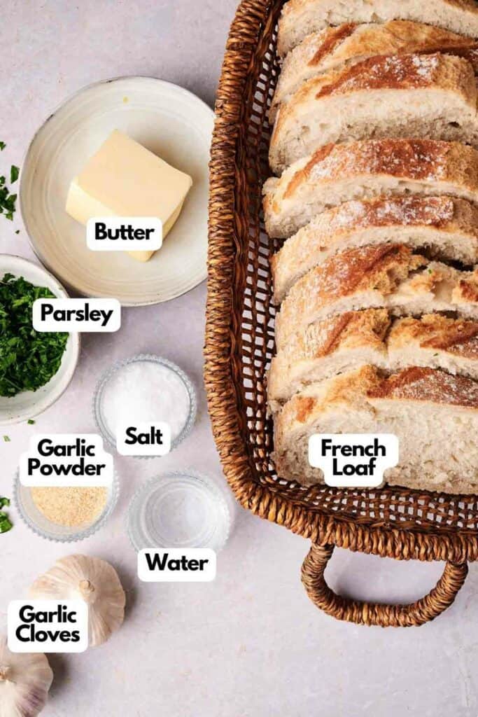 A wicker basket contains slices of French loaf. Nearby are labeled ingredients: a plate of butter, chopped parsley, a bowl of garlic powder, salt, water, and two garlic cloves&mdash;everything you need for perfect garlic bread.