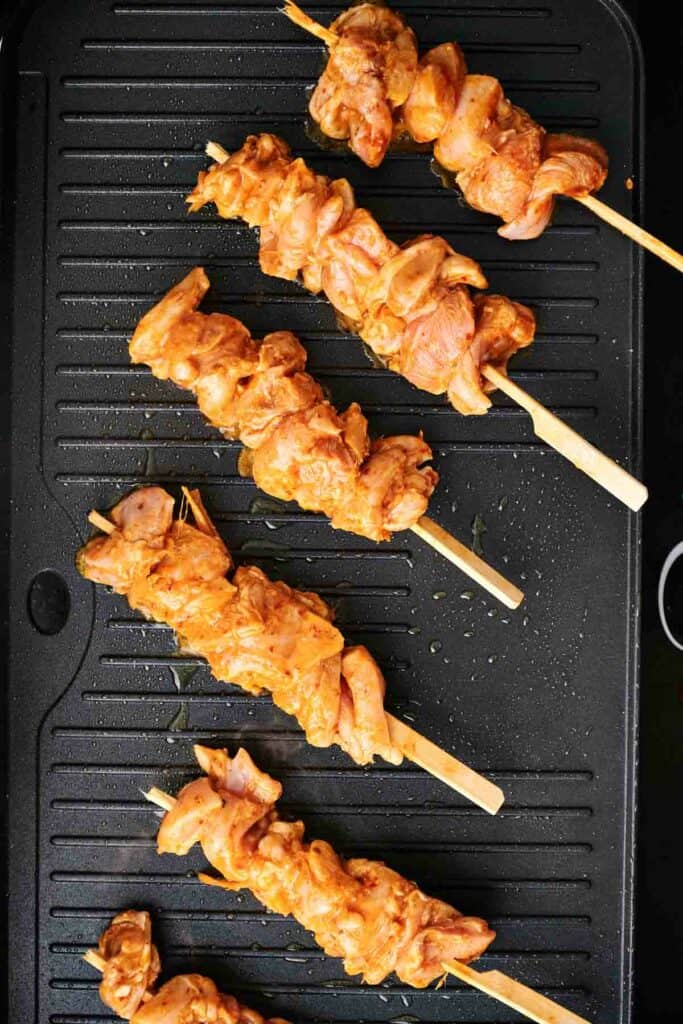 Five skewers of seasoned Thai chicken are being grilled on a black stovetop grill.
