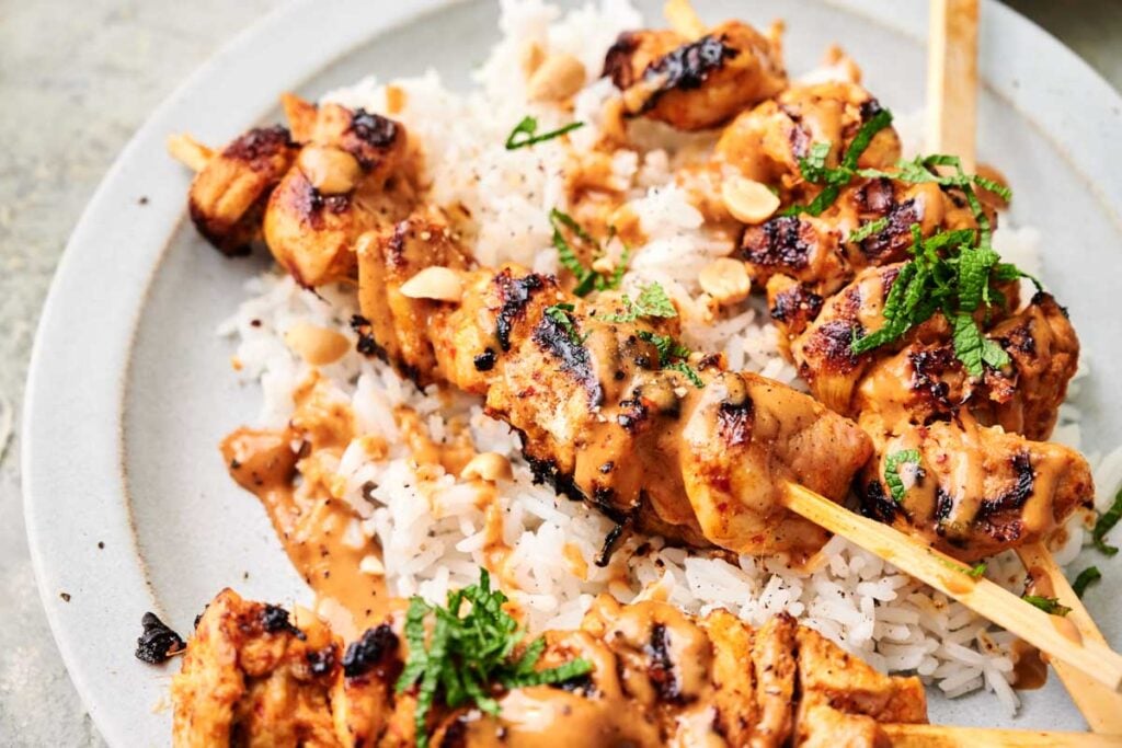 Thai chicken skewers grilled to perfection with peanut sauce, served over white rice and garnished with chopped peanuts and fresh herbs on a white plate.