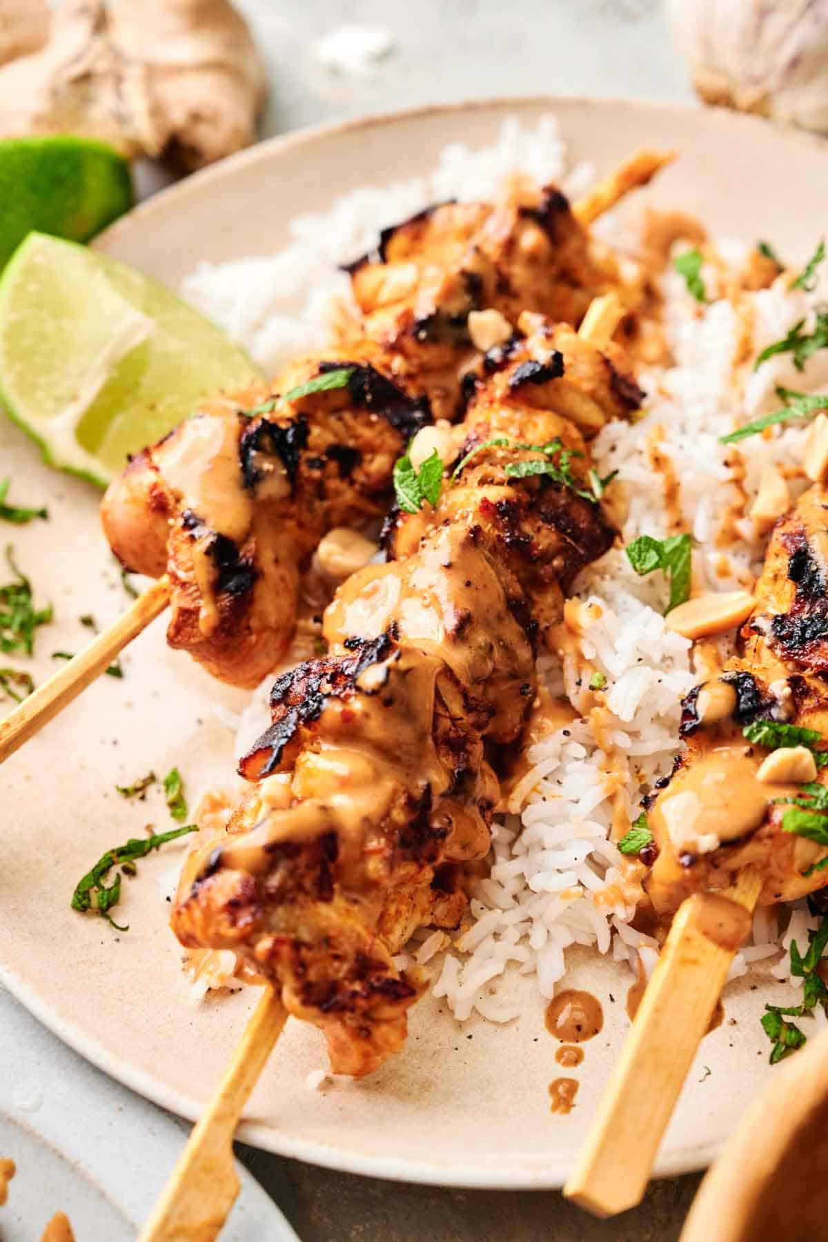 Close-up of Thai chicken skewers on a bed of white rice, garnished with chopped herbs and served with lime wedges.