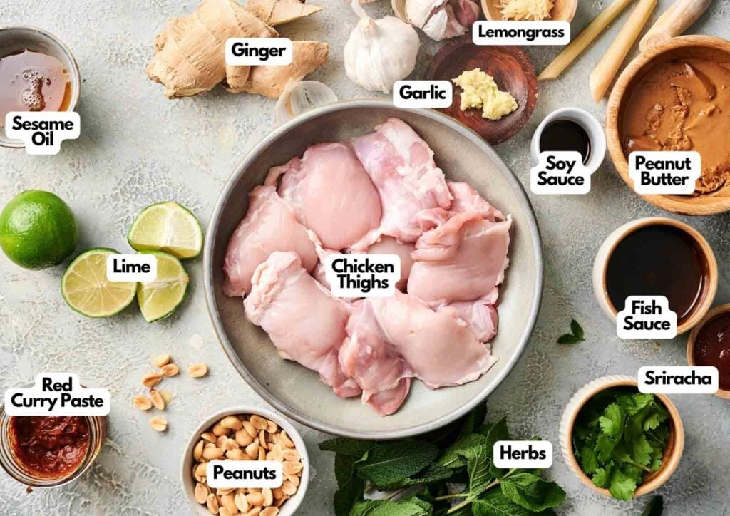 Raw chicken thighs in a bowl surrounded by labeled ingredients for Thai chicken skewers: ginger, lemongrass, garlic, soy sauce, peanut butter, fish sauce, sriracha, herbs, peanuts, red curry paste, lime, and sesame oil.
