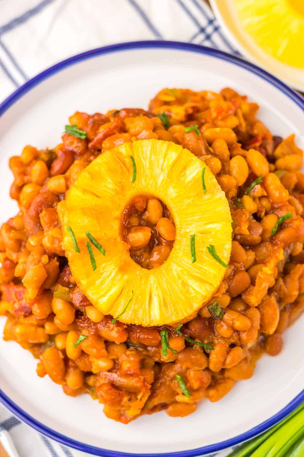A plate of Hawaiian baked beans in a tomato-based sauce, topped with a slice of pineapple and garnished with green herbs.