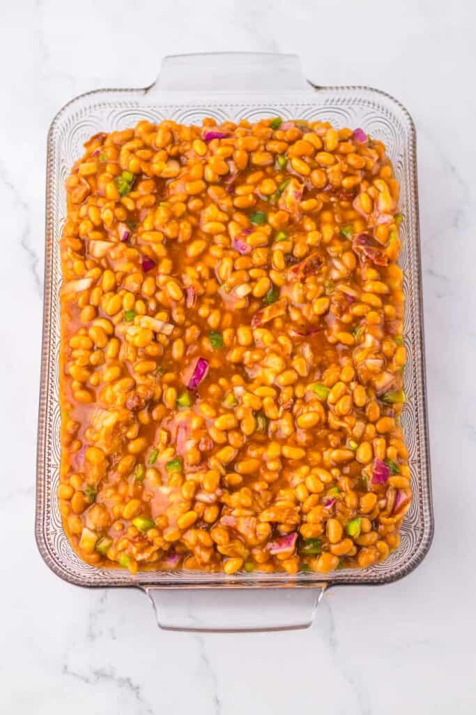 A clear glass baking dish filled with Hawaiian baked beans, mixed with pieces of red onion and green pepper, sits invitingly on a marble surface.