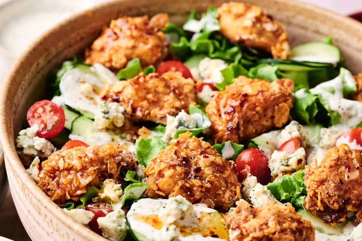 A bowl of honey hot chicken salad contains fried chicken pieces, mixed greens, cherry tomatoes, cucumber slices, blue cheese crumbles, and dressing.