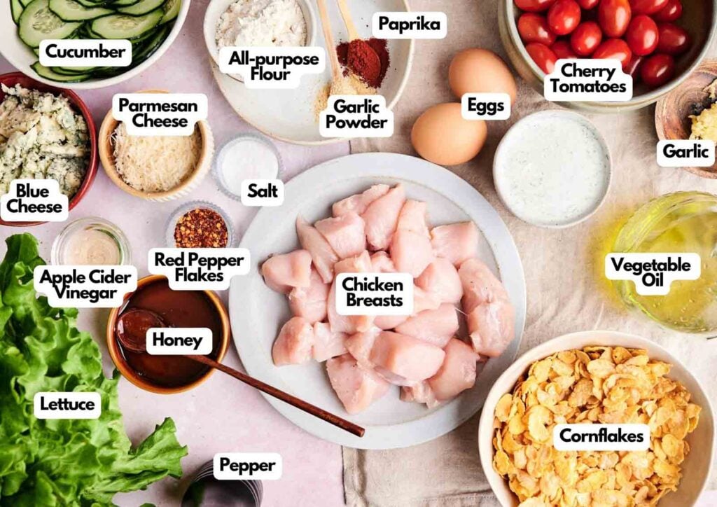 Various labeled ingredients for a hot honey chicken salad recipe, including chicken breasts, eggs, cheese, vegetables, spices, oil, cornflakes, flour, and vinegar, arranged on a tabletop.