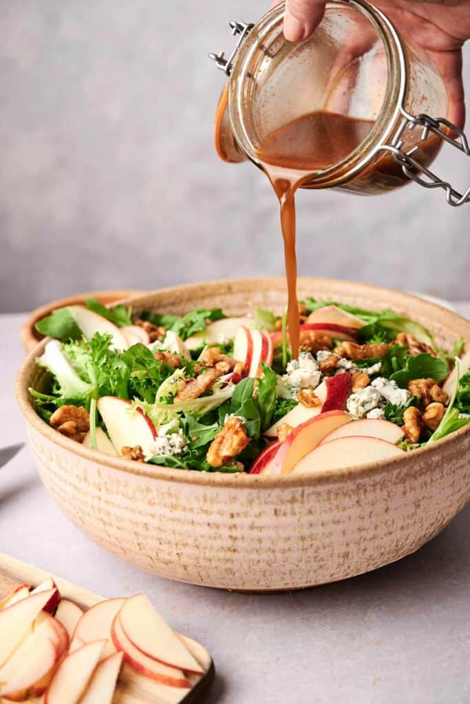 A hand pours dressing onto a honeycrisp salad in a ceramic bowl filled with greens, apple slices, walnuts, and blue cheese. A cutting board with more apple slices is visible in the foreground.