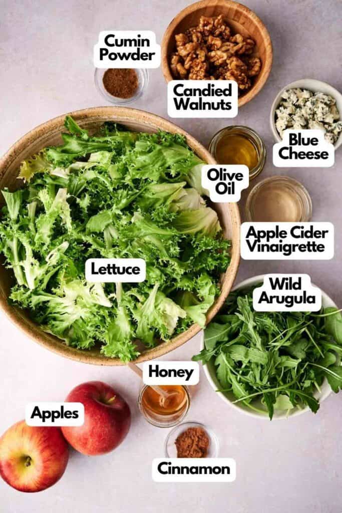 A flat lay of Honeycrisp salad ingredients including lettuce, wild arugula, candied walnuts, blue cheese, apple cider vinaigrette, cumin powder, honey, olive oil, apples, and cinnamon, all labeled.