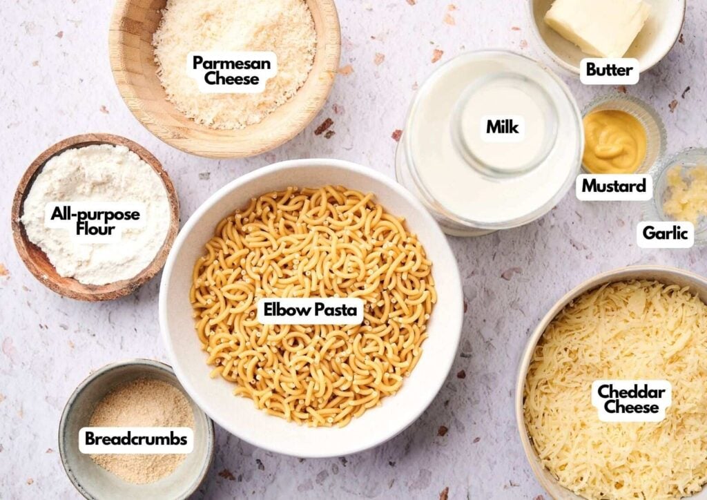 Bowls and containers filled with elbow pasta, parmesan cheese, cheddar cheese, milk, butter, mustard, garlic, all-purpose flour, and breadcrumbs arranged on a light surface—perfect ingredients for homemade mac and cheese.