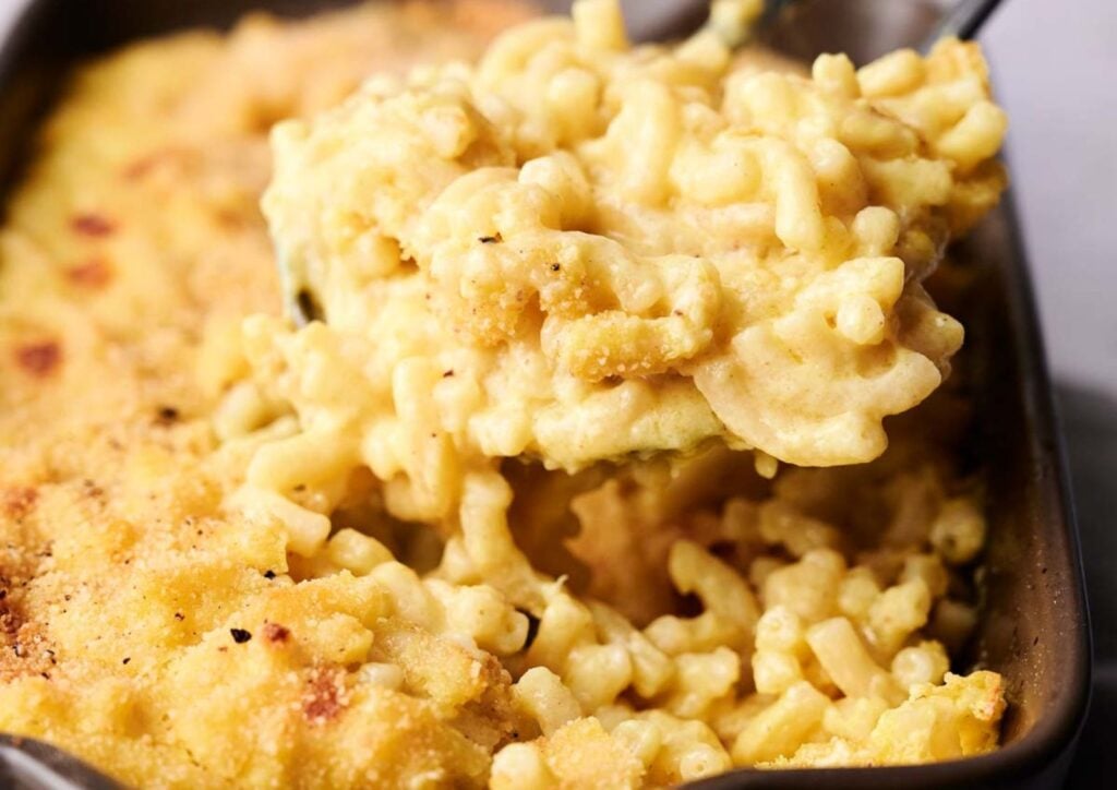 A close-up of mac and cheese being scooped from a casserole dish, showing creamy pasta with a golden breadcrumb topping.