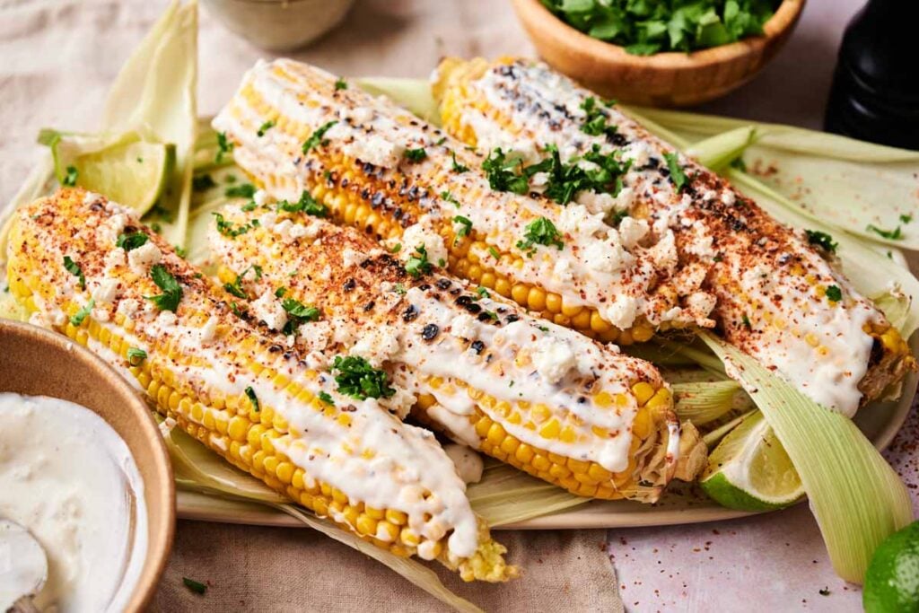 Mexican Street Corn: Grilled corn on the cob topped with creamy sauce, crumbled cheese, chili powder, and chopped cilantro, served with lime wedges and a bowl of sauce.