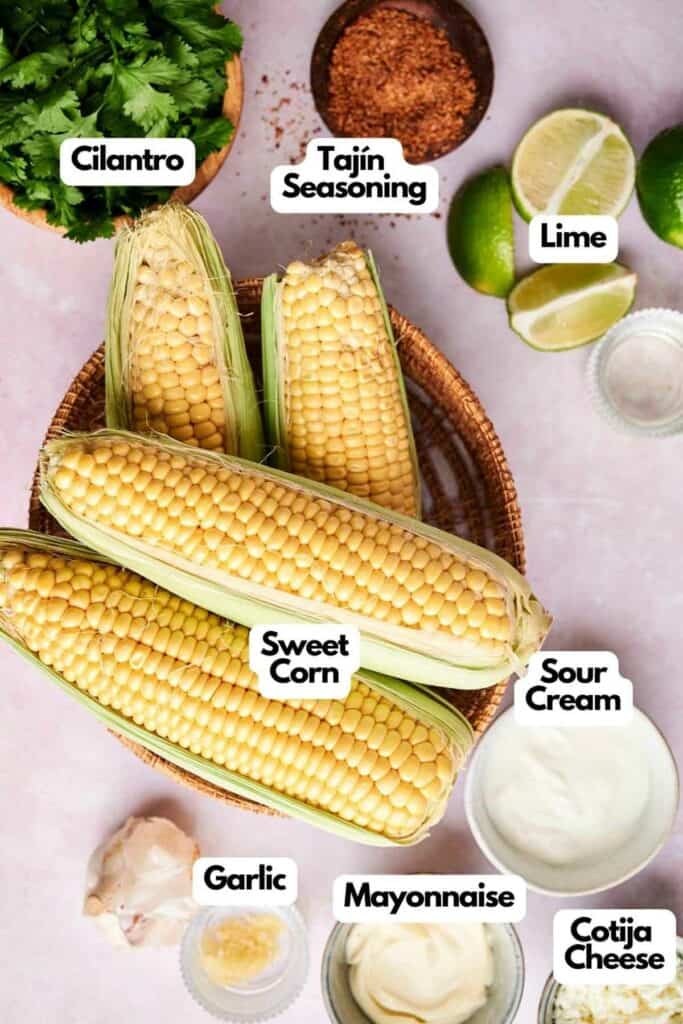 Top view of ingredients labeled around Mexican Street Corn: cilantro, Taj&iacute;n seasoning, lime, sour cream, mayonnaise, garlic, and cotija cheese.