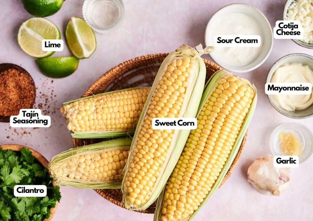 Top-down view of fresh corn in a basket surrounded by labeled ingredients for Mexican Street Corn: lime, sour cream, cotija cheese, mayonnaise, garlic, cilantro, and Tajín seasoning.