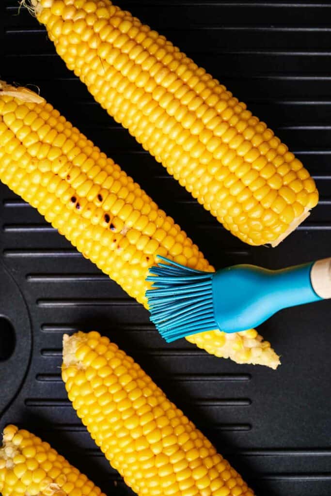 Three ears of Mexican Street Corn on the cob being brushed with a blue basting brush on a black surface.