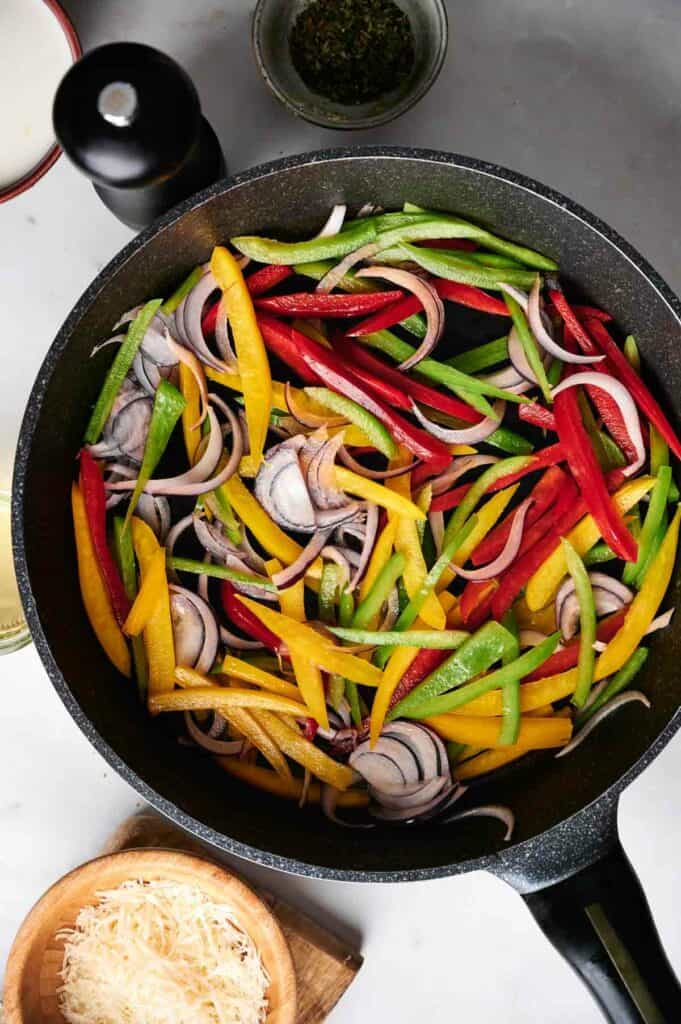 A skillet filled with cooked chicken scampi pasta mixed with bell peppers, onions, and herbs sits next to a bowl of grated cheese and a pepper grinder on a marble surface.