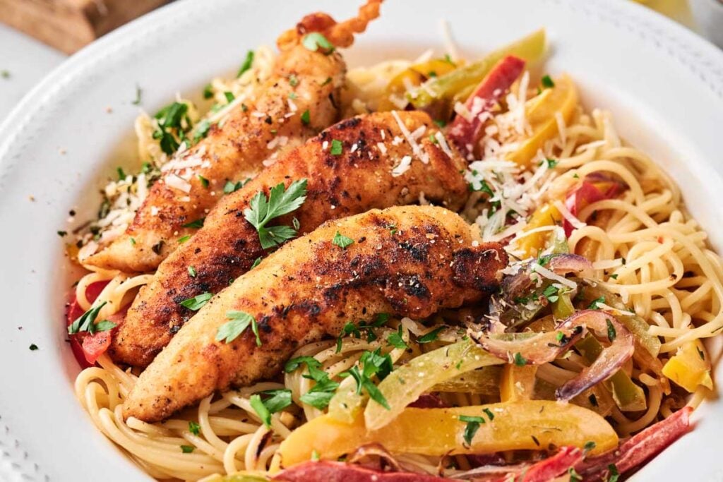 A plate of chicken scampi pasta topped with three pieces of seasoned, grilled chicken strips, sliced bell peppers, onions, and garnished with parsley and grated cheese.