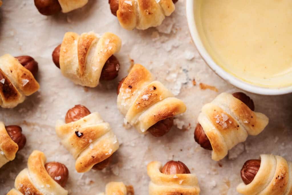 Close-up of small air fryer pigs in a blanket on a baking sheet, served with a bowl of yellow dipping sauce.