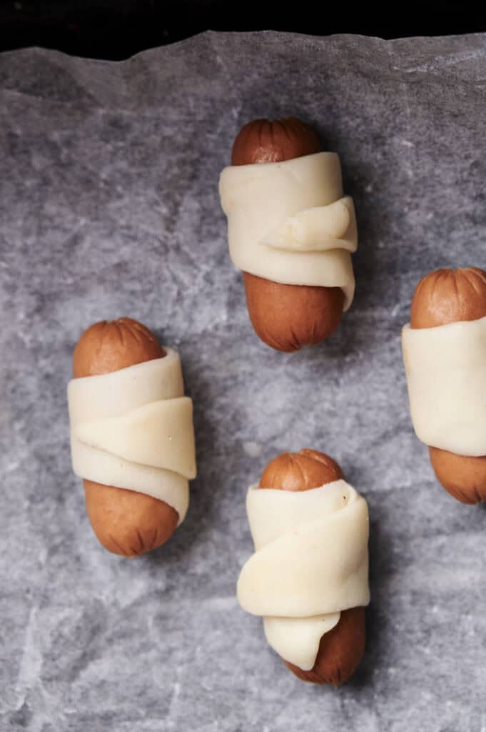 Four mini sausages wrapped in dough are placed on a parchment paper-lined baking sheet, ready to be cooked as air fryer pigs in a blanket.