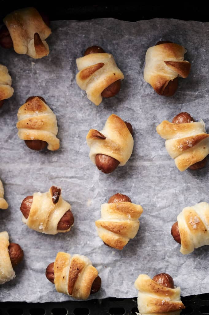 Air fryer pigs in a blanket: small sausages wrapped in golden-brown pastry, baking to perfection on a parchment-lined tray.