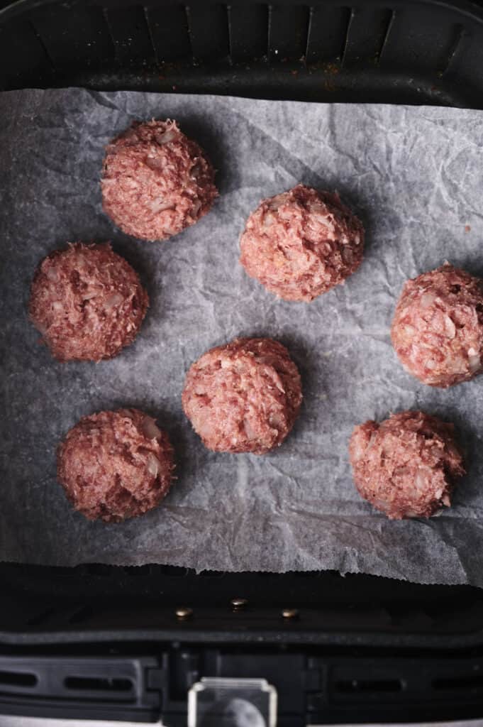 Seven raw ground meatballs on parchment paper inside an air fryer basket.