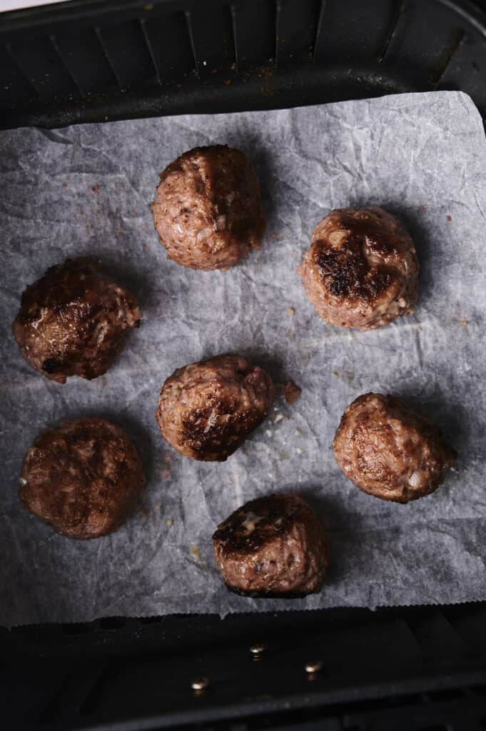 Seven cooked meatballs on parchment paper in a black air fryer basket.