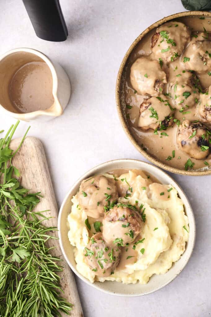 Two bowls of creamy mashed potatoes topped with gravy and meatballs. A wooden board with fresh herbs and a small jug of gravy are also visible.