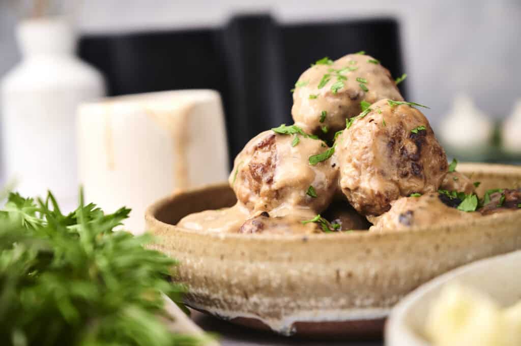 A ceramic bowl filled with meatballs covered in creamy sauce and garnished with green herbs. A small plant is partially visible in the foreground, and blurred kitchen items are in the background.