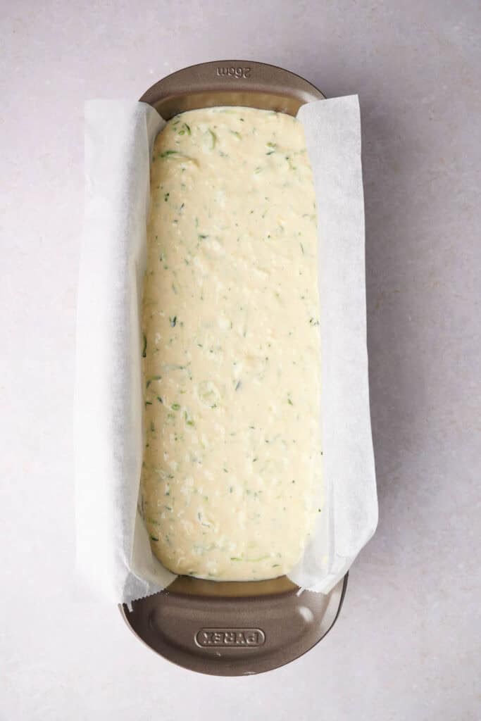Baking tin lined with parchment paper filled with uncooked batter, featuring visible green herbs, ready to be baked.