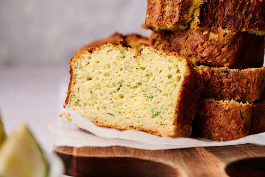 Close-up of thick zucchini bread slices stacked on a wooden cutting board, revealing a moist, textured crumb with green flecks.