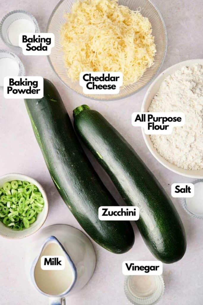 Ingredients for a recipe are laid out: two zucchinis, cheddar cheese, all-purpose flour, baking powder, baking soda, salt, milk, vinegar, and chopped green onions. Each item is labeled.